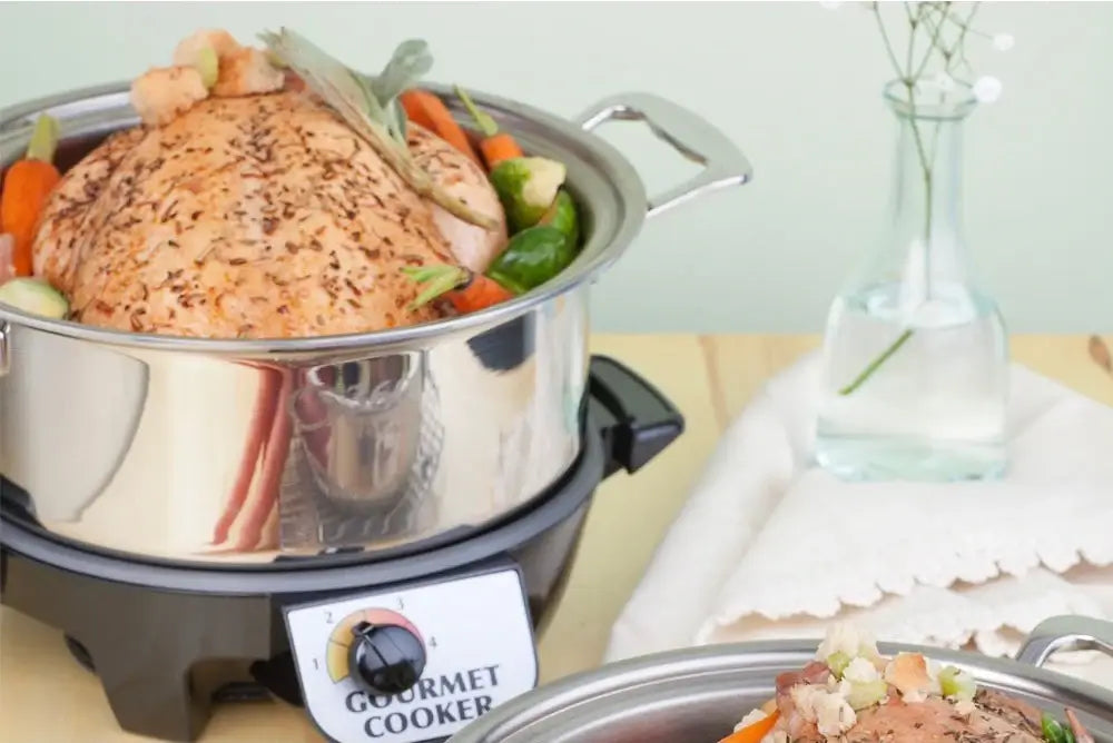 A stainless steel slow cooker filled with vegetables and a whole chicken, with a vase of flowers in the background.
