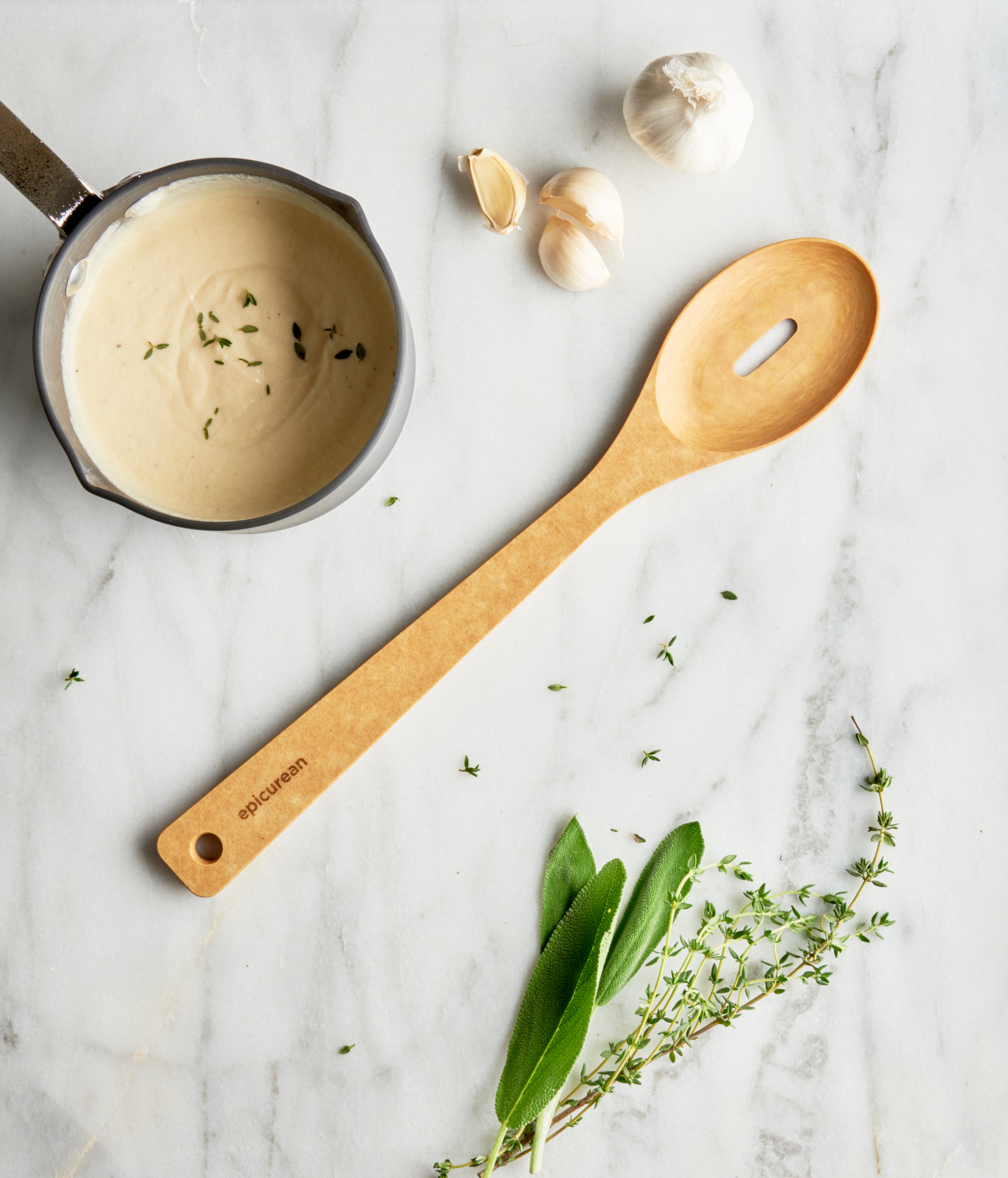 Natural slotted spoon on a marble surface with garlic and herbs, next to a pot of sauce